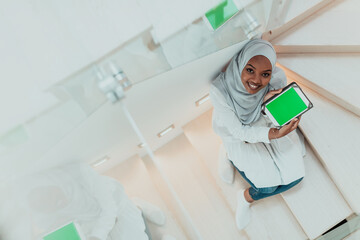 Young african modern muslim woman using tablet computer while sitting on the stairs at home wearing hijab clothes top view