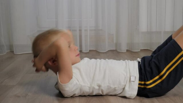 A little eight-year-old boy in shorts and a T-shirt shakes his abs lying on the floor at home. Training at home during quarantine.