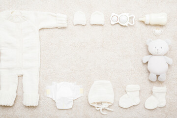 Newborn white knitted warm overalls, hat, mittens, socks, diaper, milk bottle, soother, teddy bear and rattle toy on light carpet background. Baby goods set. Empty place for text. Top down view.