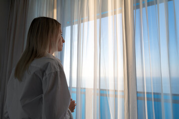 Portrait of a thoughtful young blonde woman in a white shirt at the window looking at the sea through a white curtain