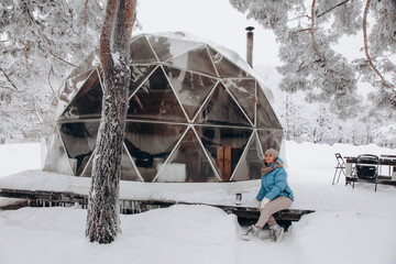A woman sits and enjoys the view next to a dome tent in glamping in the winter forest - obrazy, fototapety, plakaty