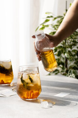 A woman pours iced tea from a bottle into an ice glass by a sunny window 