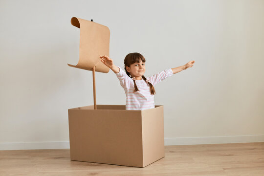 Portrait of happy positive dark haired little girl playing in handmade ship from cardboard box with sail, posing with raised arms, imagines she is sailing.