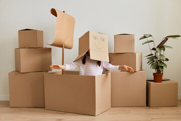 Portrait of anonymous little girl sitting in cardboard ship with sail with carton box with drawn face on her head, spreading hands asides, having fun while relocating.