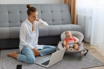 Horizontal shot of tired oman in white shirt and jeans sitting on floor near sofa with baby in...