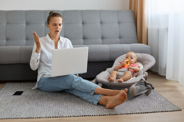 Full length portrait of confused shocked woman wearing white shirt and jeans sitting on floor near sofa with baby in rocking chair, female freelancer looking at screen with puzzlement.