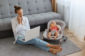 Indoor shot of busy Caucasian woman wearing white shirt and jeans sitting on floor near sofa with baby daughter in rocking chair, using laptop while talking phone.