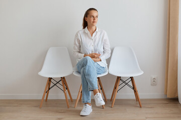 Portrait of sick ill woman wearing white shirt and jeans sitting on chair in queue to the doctor,...