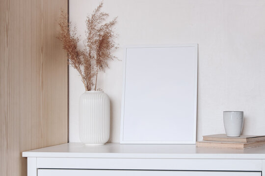 Blank photo frame and dry reed in a white vase, a cup of coffee on an old book. Modern Scandinavian style interior on the beautiful chest of drawers with white wall and bright room. Elegant interior