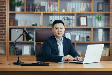 Portrait of a successful Asian businessman, man working in the office sitting at the table, looking at the camera and smiling, happy banker with laptop