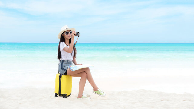 Happy traveler and tourism young women travel summer on the beach. Asian smiling people holding smartphone take photo  and sitting on yellow suitcase for relax outdoor destination leisure trip travel