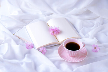Obraz na płótnie Canvas 白い布の上のピンクのコーヒーカップのコーヒーと八重桜の花と見開きの本