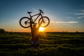 man stand in action lifting bicycle above his head on the meadow with sunset
