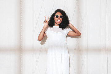 Beautiful black woman with afro curls hairstyle. Smiling model dressed in white summer dress. Sexy carefree female posing near wall in studio. Tanned and cheerful. At sunny day. Shadow from window