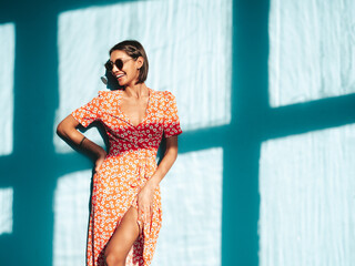 Young beautiful smiling female in trendy summer red dress. Sexy carefree woman posing near blue wall in studio. Positive model having fun. Cheerful and happy. At sunny day. Shadow from window