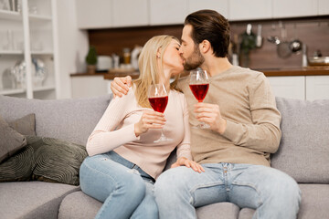 Happy marriage anniversary. Cheerful spouses celebrating anniversary at home, sitting on the sofa and holding glasses with wine, enjoying time together, kissing
