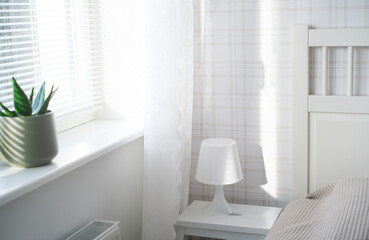 The interior of the room in white tones with a bed, bedside table, table lamp and a flower on the windowsill