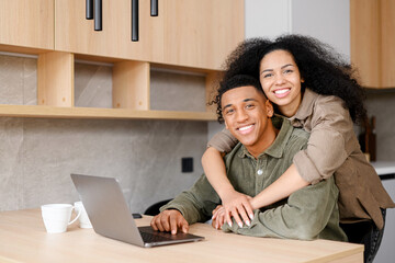 Biracial woman hugging man from behind. Mixed ethnicity couple relaxing in new apartment at home while browsing internet on laptop. Show love everyday in the simplest of ways concept