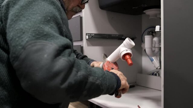 Mature plumber cutting the pipe to install sink siphon in domestic kitchen using pvc pipe cutter. Repairman working.