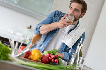 Handsome young man in casual wear drinking wine while standing in the kitchen at home.