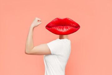 Strong woman headed by red lips raises arm and shows bicep isolated on coral color background....