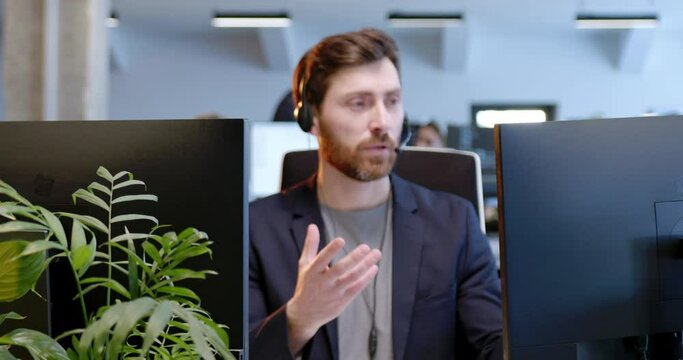 Caucasian handsome man in headset working at computer screen, talking with client online. Customer support. Male talking in headphones at call center office. Speaking with customers. Zoom in.