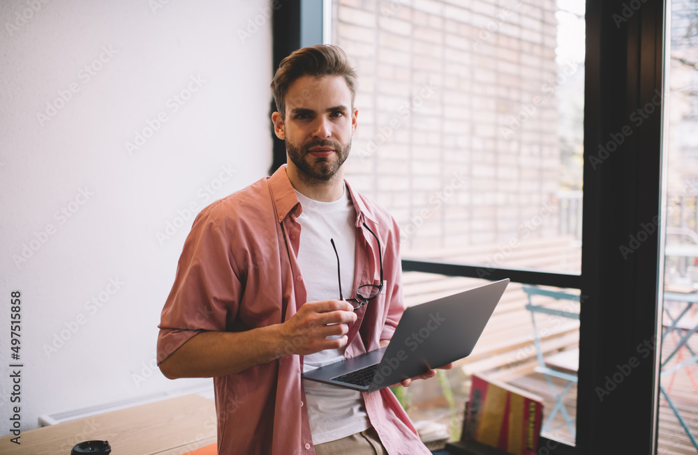 Poster portrait of skilled male freelancer with modern laptop computer for web programming looking at camer