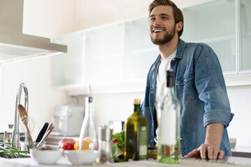 Handsome smiling young man leaning on kitchen counter with vegetables and looking at camera.