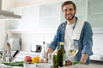 Handsome smiling young man leaning on kitchen counter with vegetables and looking at camera.