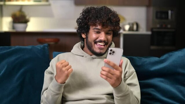 Happy satisfied Indian man holding smartphone and smiling making yes gesture, multiracial guy celebrating online lottery or giveaway victory sitting on the sofa at home