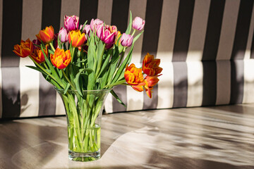 A bouquet of yellow and pink tulips in a glass vase stands on the floor in bright sunlight with contrasting shadows