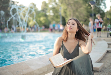 smiling young woman sitting on the edge of a fountain in a park reads a book