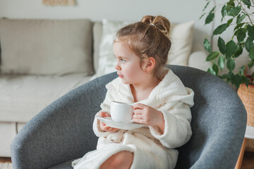 Little curly-haired girl in a soft light dressing gown drinks tea and relaxes at home sitting in a...