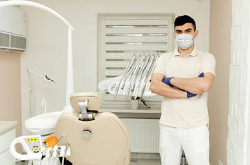 Practicing dentist in a protective mask, and in protective gloves with his hands folded on his chest while in the dental office.