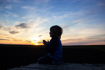 silhouette of a boy sitting on his knees, hands folded in prayer, turning to God at sunset. Children against war. the child asks for peace for his homeland Ukraine