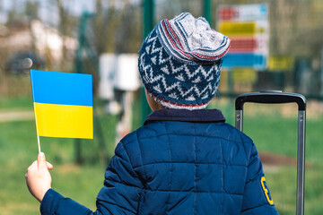 Child or kid with luggage, suitcase or bag, winter clothes, hat and Ukrainian flag during war in...