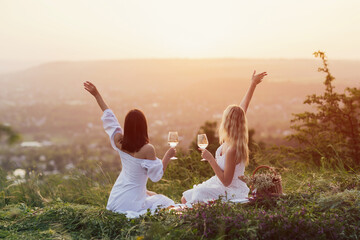 Two young happy female with hands up in white summer dresses at picnic. Carefree women making...