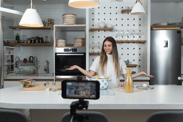 Young vlogger woman vlogging about cooking at her apartment domestic kitchen cook and making video vlog with smartphone as online content creator for internet. Female influencer making a meal.