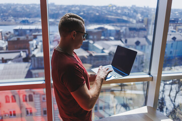 Smiling businessman stands by the window while working on a laptop. Multitasking concept. A busy freelancer in glasses holds a laptop in his hands while standing near the window. Smart casual.