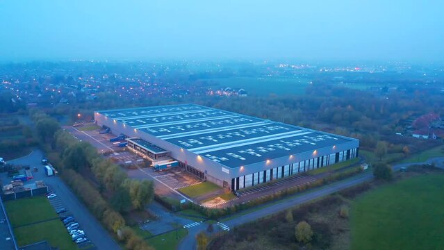Warehouse at dusk: Aerial moving back up, with the lights of the nearby city and roads. Cinematic. Wattrelos, France