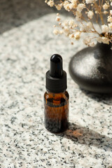 Bottle with dropper pipette with serum or essential oil. Grey granite background with daylight and yellow flowers. Skincare products, natural cosmetic. Beauty concept for face and body care