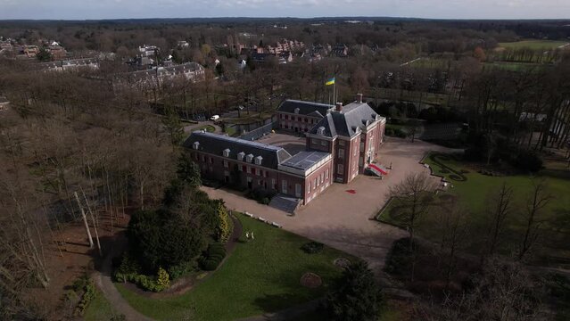 Descending aerial showing Slot Zeist castle between trees with the moated manor surrounded by green park and urban landscape in the background. Dutch stately venue seen from above.
