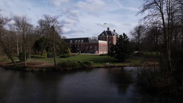 Sideways aerial over pond in front of Slot Zeist castle with the moated manor surrounded by green park and urban landscape in the background. Ukrainian flag on Dutch stately venue seen from above.