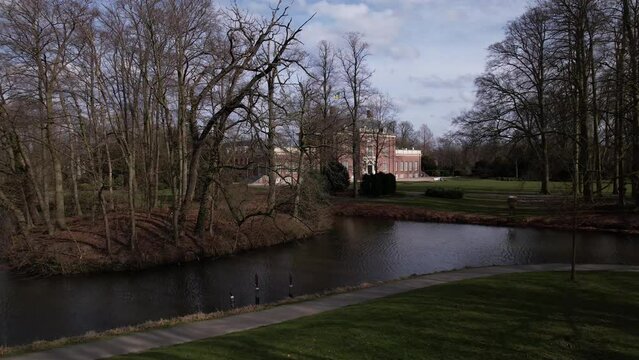 Ascending aerial revealing Slot Zeist castle behind winter barren trees with the moated manor surrounded by green park and urban landscape in the background. Dutch stately venue seen from above.