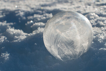 A single crystalline translucent thin shelled ice bubble frozen with frosty patterns and particles in the globe. The round soap bubble is on white snow with crystal patterns in the icy frost ball.