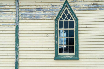 The exterior wall of a religious building with pale yellow colored narrow horizontal clapboard siding. There's a tall Gothic style window with clear glass, white wooden muntins, and green wood trim. 