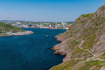 A footpath, hiking trail, or path along a hillside. The cliff is rocky with grass patches. The city of St. John's, Newfoundland, is in the background on a sunny day. The sky is bright blue. 