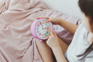 Toddler girl, whose face is not visible, sits in bed and holds an alarm clock in her hands, learns...