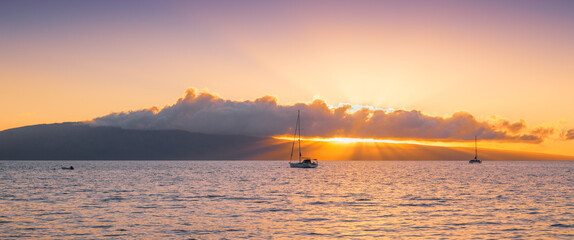 Rays of light from colorful sunset beneath clouds over an island and sailboats - panorama view of...