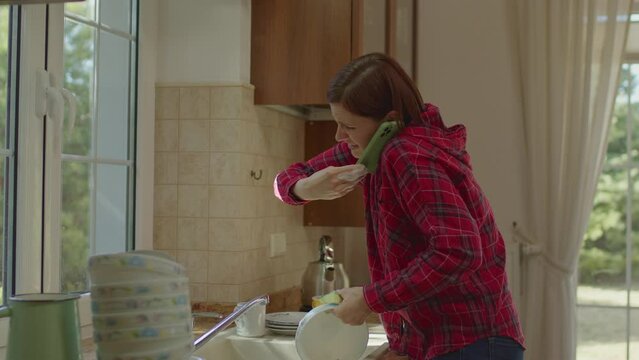 Woman talking on the mobile phone while dishwashing in the kitchen. Using cell phone with nose because of dirty hands.